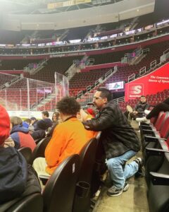 man talking to a younger man who is sitting in front of him in a stadium- photo from profile